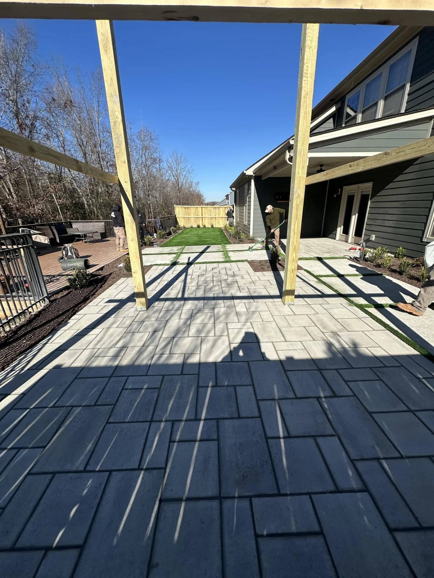 Workers landscape a modern backyard patio beneath pergola framing.