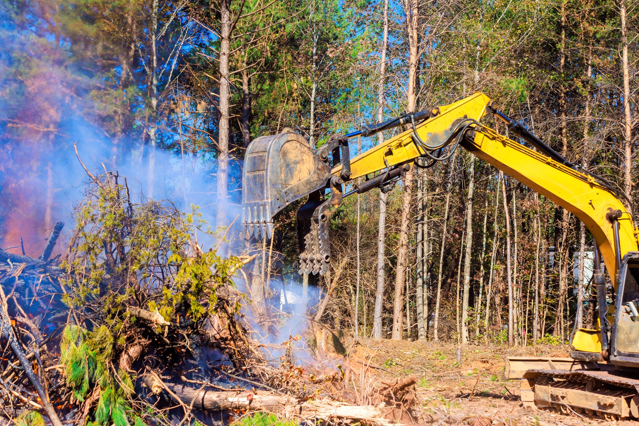 Excavator clears burning brush pile in wooded area