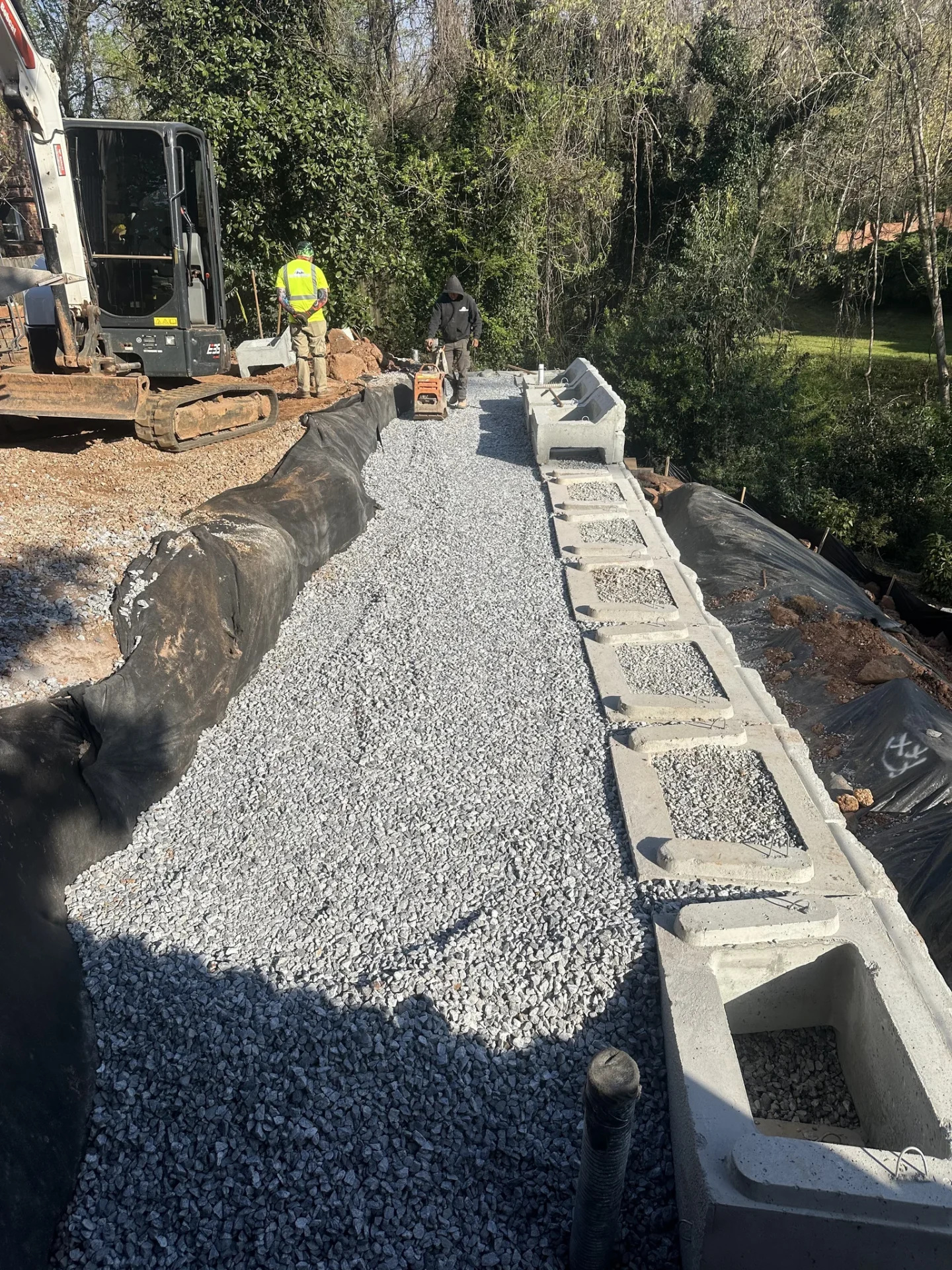 Workers compact gravel beside excavator and concrete drainage channel
